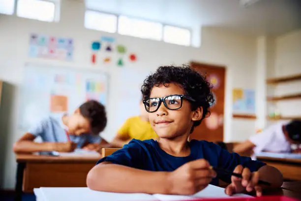 Schoolboy wearing eyeglasses sitting at desk, looking sideways, smiling in class